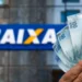 Mão segurando várias notas de 100 reais com fundo desfocado do logo da Caixa Econômica Federal