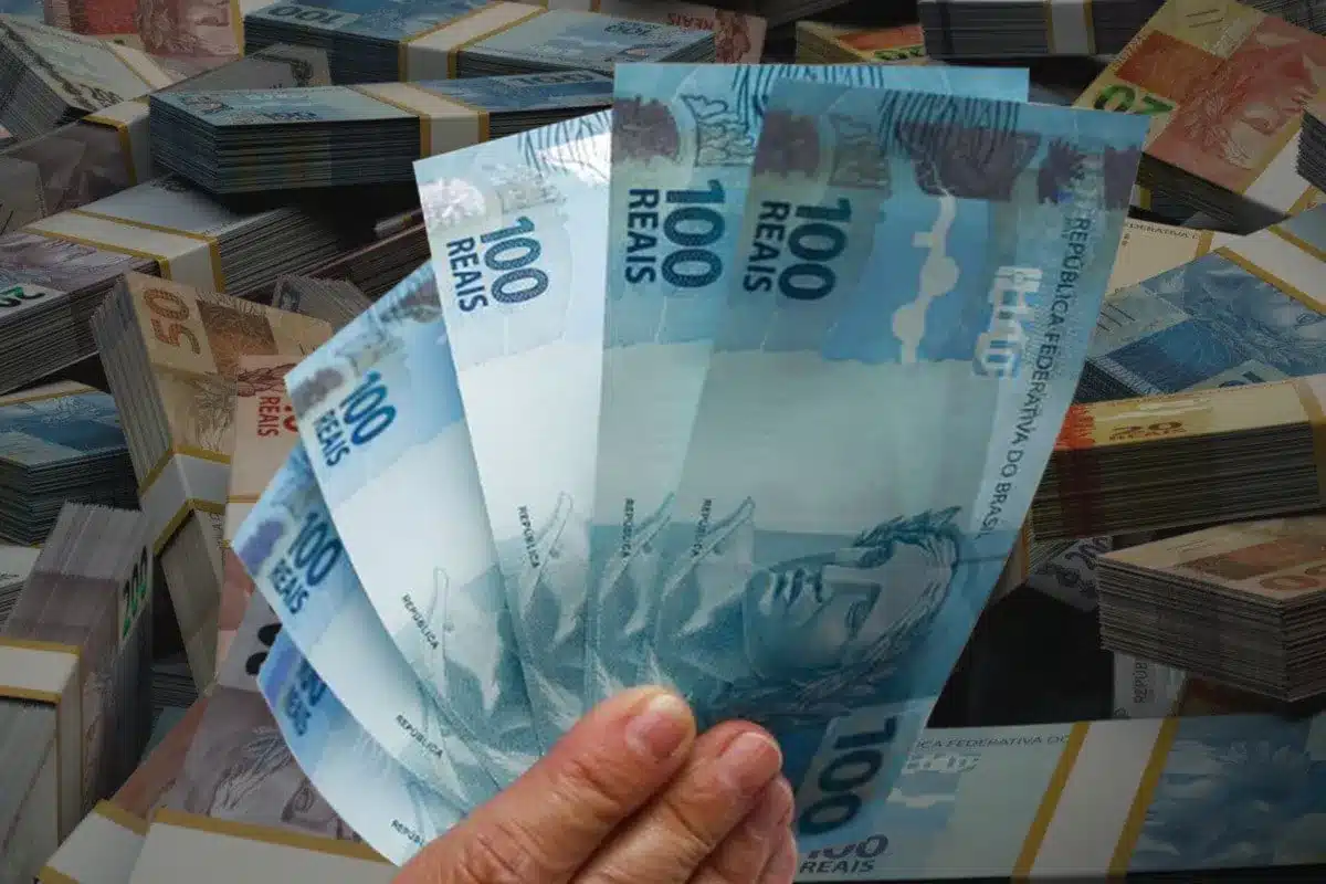 13 Salário Parcelas Datas Pagamento INSS Notas de R$ 100 e moedas de Real. A imagem representa o pagamento do 13º salário, que é feito em duas parcelas, com descontos de INSS e Imposto de Renda.