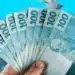 Mão segurando um leque de notas de R$ 100, com mais dinheiro caindo sobre um fundo azul, simbolizando um abono extra ou saque de valores.