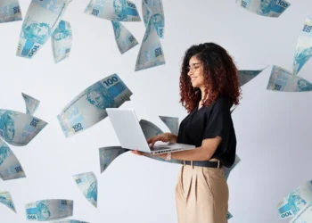 Mulher sorridente usando notebook com várias cédulas de 100 reais caindo ao fundo