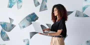 Mulher sorridente usando notebook com várias cédulas de 100 reais caindo ao fundo