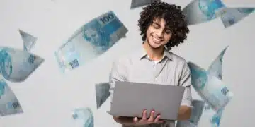Homem jovem sorridente usando laptop com cédulas de 100 reais voando ao redor