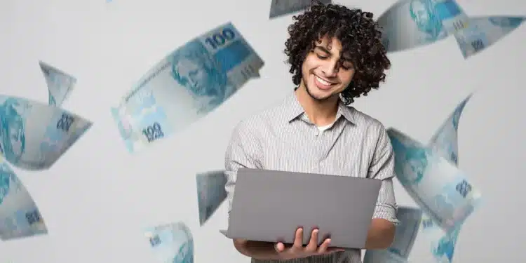 Homem jovem sorridente usando laptop com cédulas de 100 reais voando ao redor
