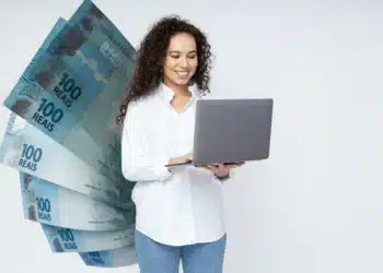 Mulher sorridente com notebook e notas de 100 reais ao fundo, representando a gestão do 13º salário.