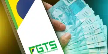 Mão segurando notas de 100 reais ao lado de um celular com o logo do FGTS, representando a antecipação do Saque-Aniversário e a nova regra de acesso ao fundo de garantia