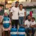 Família sorridente com botijões de "Gás do Povo" em uma comunidade, representando o benefício social para famílias do Bolsa Família.