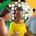 Três idosos sorrindo e conversando em ambiente externo com decoração verde e amarela ao fundo