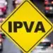 Placa amarela com a sigla IPVA em destaque sobre tráfego de carros em uma rua movimentada