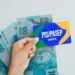 Mão segurando o cartão PIS/PASEP da CAIXA, com notas de R$ 100 ao fundo, ilustrando o saque de até R$ 2.800 disponível até 30 de novembro.