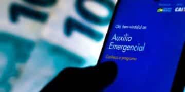 Mão segurando smartphone com o aplicativo "Auxílio Emergencial" na tela e notas de dinheiro ao fundo, representando pagamentos ou débitos.