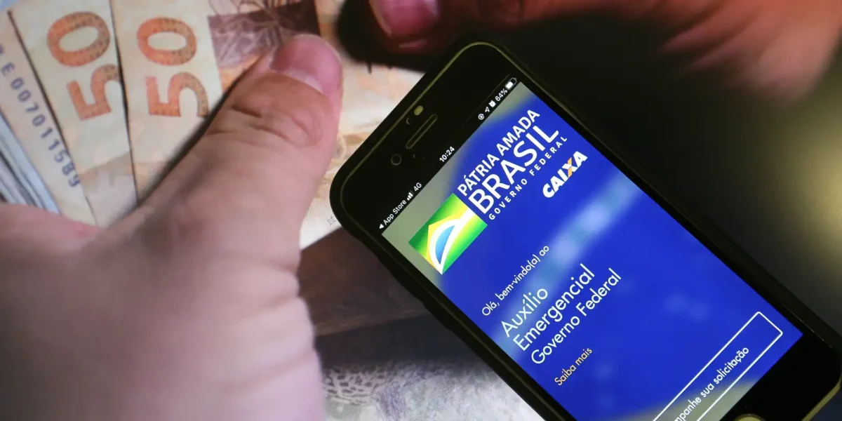 Ressarcimento do Auxílio Emergencial Mão segurando notas de cinquenta reais ao lado de um celular exibindo a tela inicial do Auxílio Emergencial.