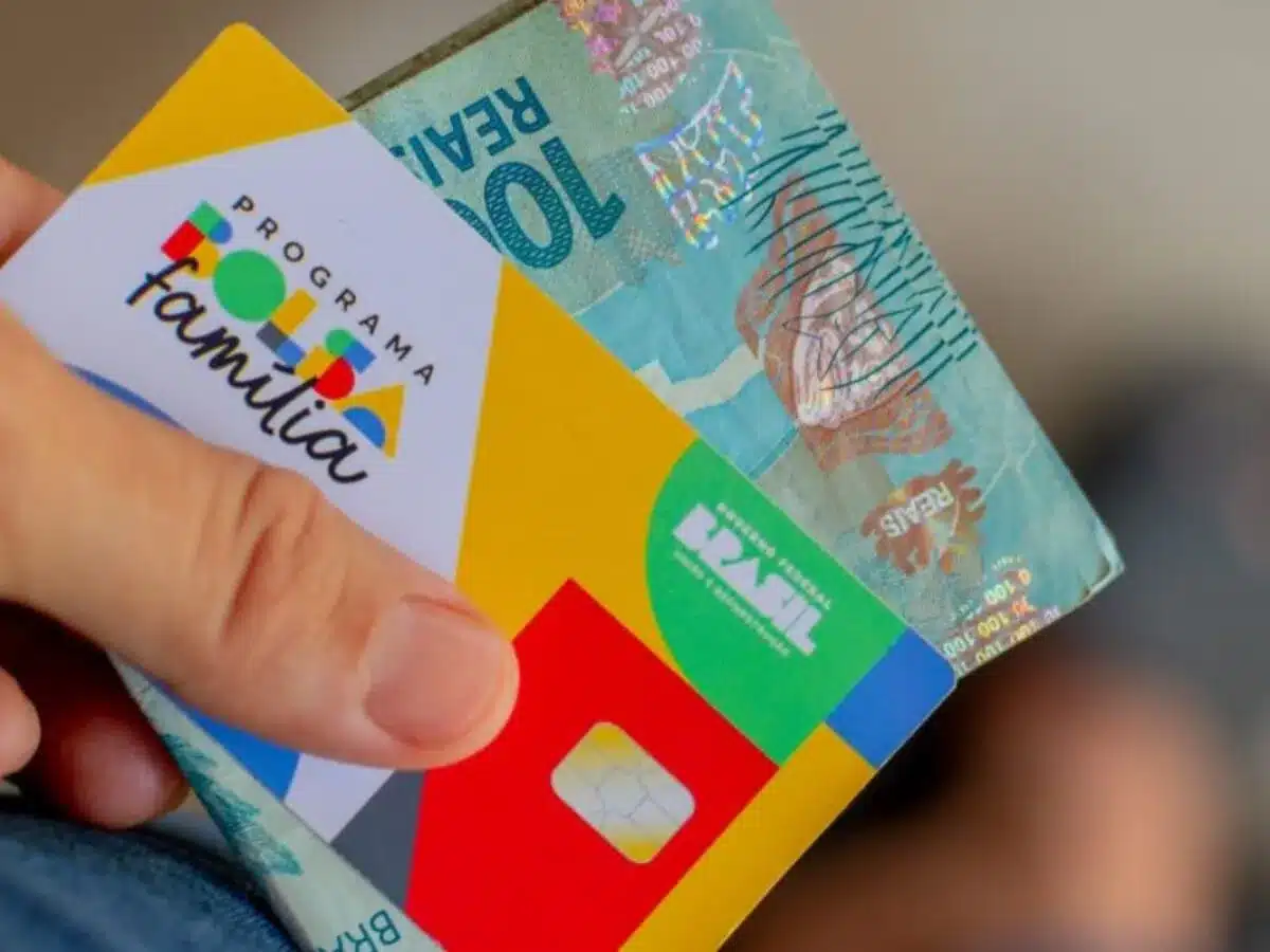 Calendário Oficial do Bolsa Família Dezembro 2025 e Valor Médio do Benefício Mão segurando o cartão do Programa Bolsa Família e uma nota de 100 Reais. Imagem ilustrativa do depósito do benefício social de dezembro, pago entre os dias 10 e 23.