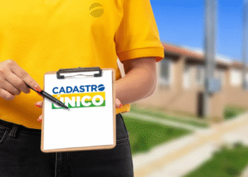 Pessoa com camisa amarela segurando prancheta com logotipo "Cadastro Único" e apontando com caneta, em fundo externo desfocado.