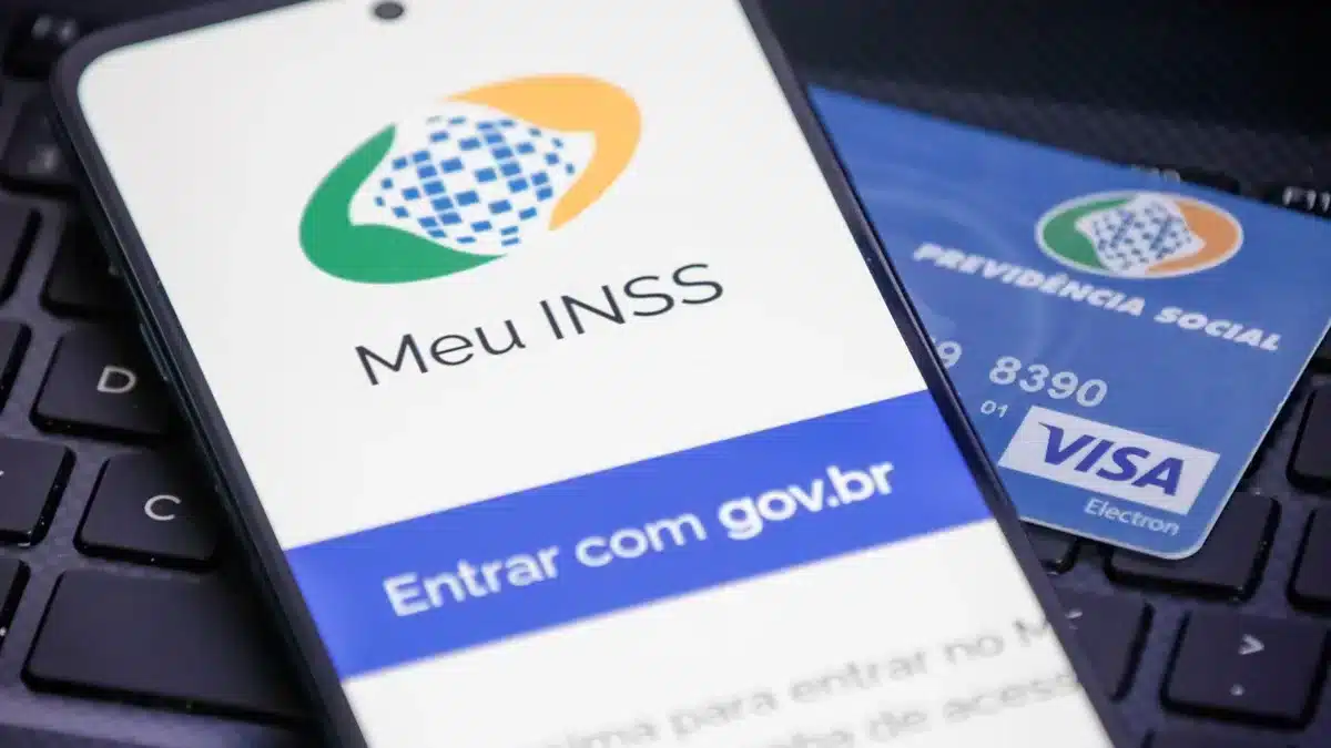 Meu INSS: Sua Porta de Entrada para a Restituição de Descontos Indevidos Imagem aproximada de um celular exibindo o aplicativo "Meu INSS" sobre um teclado de notebook, com um cartão da Previdência Social ao fundo, simbolizando o acesso digital aos benefícios e a restituição de descontos.