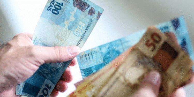 Pessoa segurando notas de reais brasileiras (R$ 100 e R$ 50) enquanto calcula o valor do 13º salário.