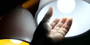 Mão próxima a uma lâmpada acesa representando o aumento no consumo de energia elétrica.