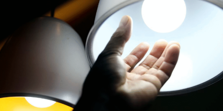 Mão próxima a uma lâmpada acesa representando o aumento no consumo de energia elétrica.