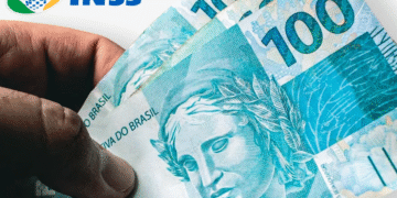 Mão segurando três notas de 100 reais com o logo do INSS ao fundo, ilustrando pagamentos de benefícios previdenciários.