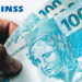 Mão segurando três notas de 100 reais com o logo do INSS ao fundo, ilustrando pagamentos de benefícios previdenciários.