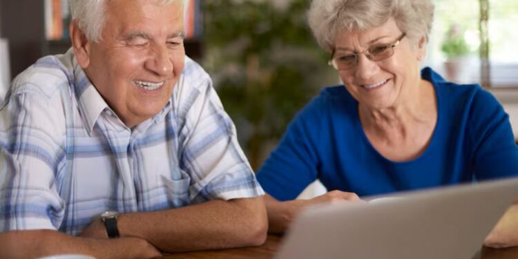 Casal de idosos felizes usando laptop em casa, representando a descoberta de um benefício de viagem.