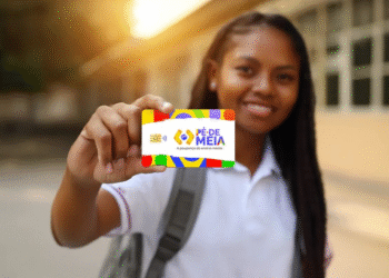 Estudante sorridente segurando o cartão colorido do programa Pé-de-Meia, com logotipo e texto "A poupança do ensino médio", em fundo desfocado de ambiente escolar ao entardecer.