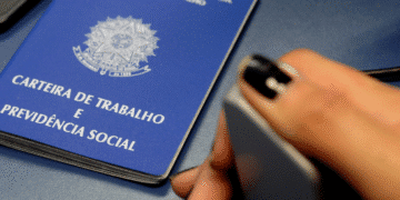 Carteira de Trabalho azul ao lado da mão de uma pessoa segurando um cartão.