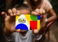 Criança segurando cartão do Programa Bolsa Família colorido em foco com adulto ao fundo