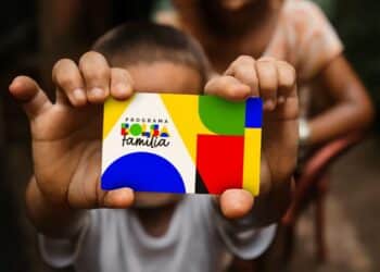 Criança segurando cartão do Programa Bolsa Família colorido em foco com adulto ao fundo