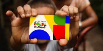 Criança segurando cartão do Programa Bolsa Família colorido em foco com adulto ao fundo