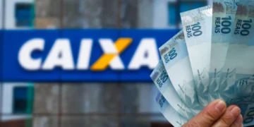 Mão segurando várias notas de 100 reais com fachada da Caixa Econômica Federal ao fundo desfocada