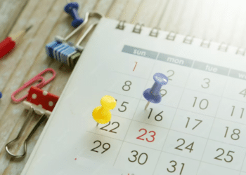 Close-up de um calendário de mesa branco com tachinhas coloridas (amarela e roxa) marcando datas, cercado por clipes de papel e um lápis sobre uma mesa de madeira.