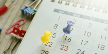 Close-up de um calendário de mesa branco com tachinhas coloridas (amarela e roxa) marcando datas, cercado por clipes de papel e um lápis sobre uma mesa de madeira.