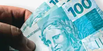 Mão segurando três cédulas de 100 reais, com foco na Efígie da República e detalhes da nota de Real.