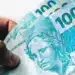 Mão segurando três cédulas de 100 reais, com foco na Efígie da República e detalhes da nota de Real.
