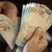 Mãos segurando notas de 50 reais, representando o depósito do PIX de R$ 710 no Caixa Tem. A imagem simboliza a transferência de dinheiro e o recebimento de benefícios.