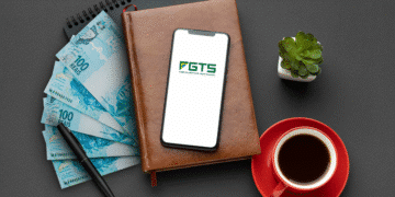 Smartphone com o logo do aplicativo FGTS (Fundo de Garantia do Tempo de Serviço) sobre um caderno de couro ao lado de notas de R$ 100 reais, uma xícara de café e uma caneta, representando a liberação do Saque-Aniversário para nascidos em dezembro de 2025.