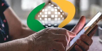 Close-up das mãos de uma pessoa idosa manuseando um celular, com o logotipo da Previdência Social centralizado na tela.