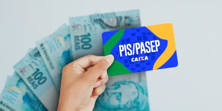 Cartão do PIS/Pasep da Caixa sendo segurado por trabalhador com cédulas de real ao fundo.