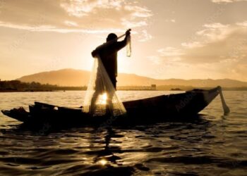 Silhueta de um pescador em pé sobre um barco de madeira no rio, preparando-se para lançar uma rede de pesca circular contra o sol do entardecer.