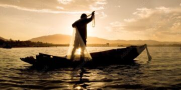 Silhueta de um pescador em pé sobre um barco de madeira no rio, preparando-se para lançar uma rede de pesca circular contra o sol do entardecer.