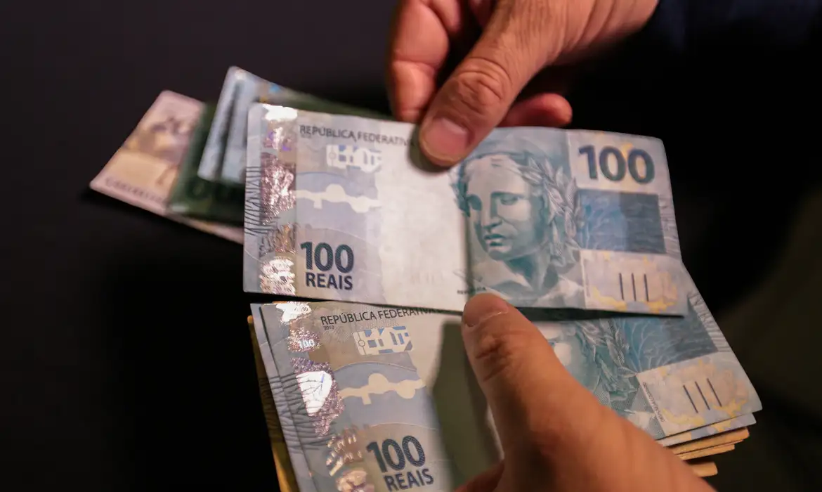 Contando cédulas de 100 reais com as mãos Mãos masculinas contando várias notas de 100 reais brasileiras sobre fundo escuro