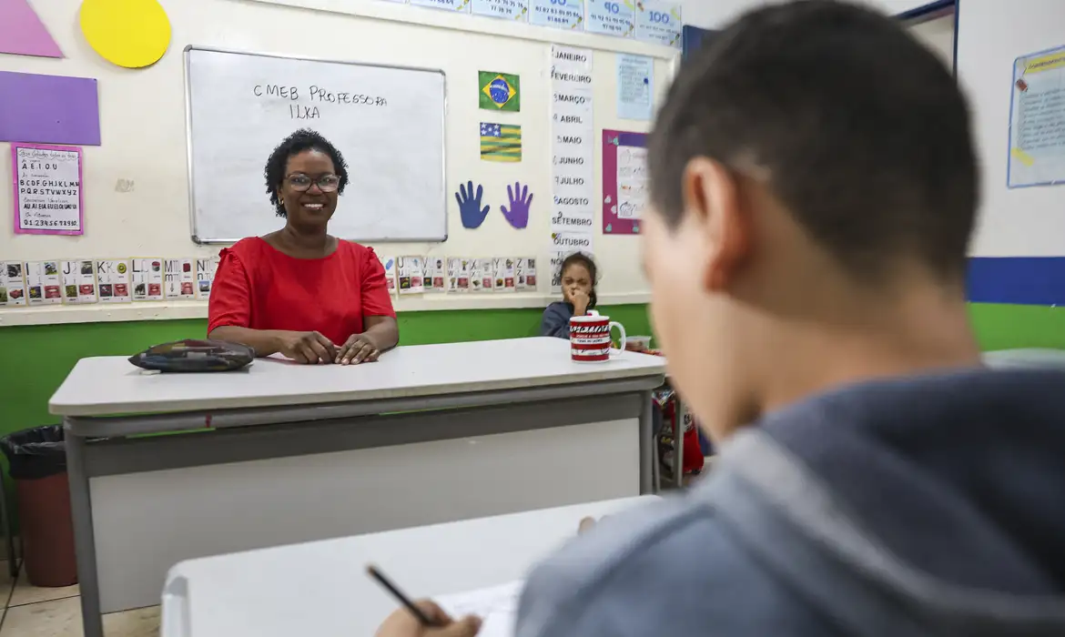 Professora sorridente em sala de aula com alunos Professora negra de óculos e camisa vermelha sentada à mesa em sala de aula com aluno escrevendo em primeiro plano