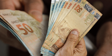 Mãos segurando um leque de notas brasileiras de 50 Reais, representando o pagamento do 13º salário do INSS.