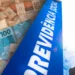 Composição visual mostrando uma placa azul vertical com a inscrição branca "PREVIDÊNCIA SOCIAL" em ângulo inclinado, sobreposta a diversas cédulas de 50 e 100 reais ao fundo.