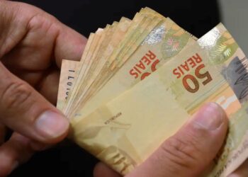 Close de mãos contando várias notas de 50 reais, representando o pagamento do imposto ou o recebimento de desconto.