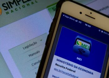 Close de um smartphone exibindo o aplicativo oficial do MEI da Receita Federal, posicionado em frente a uma tela de computador com o site do Simples Nacional ao fundo.