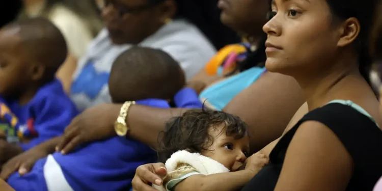Mulher amamentando bebê enquanto outras pessoas sentadas ao fundo participam da reunião