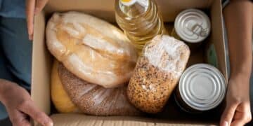 Close de mãos de voluntários organizando doações de alimentos; uma pessoa segura um saco plástico com maçãs vermelhas, outra coloca um pote de conserva em uma sacola de papel com brócolis, cercados por pães, latas e garrafas de água.