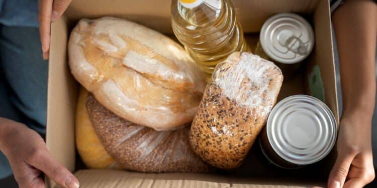 Close de mãos de voluntários organizando doações de alimentos; uma pessoa segura um saco plástico com maçãs vermelhas, outra coloca um pote de conserva em uma sacola de papel com brócolis, cercados por pães, latas e garrafas de água.