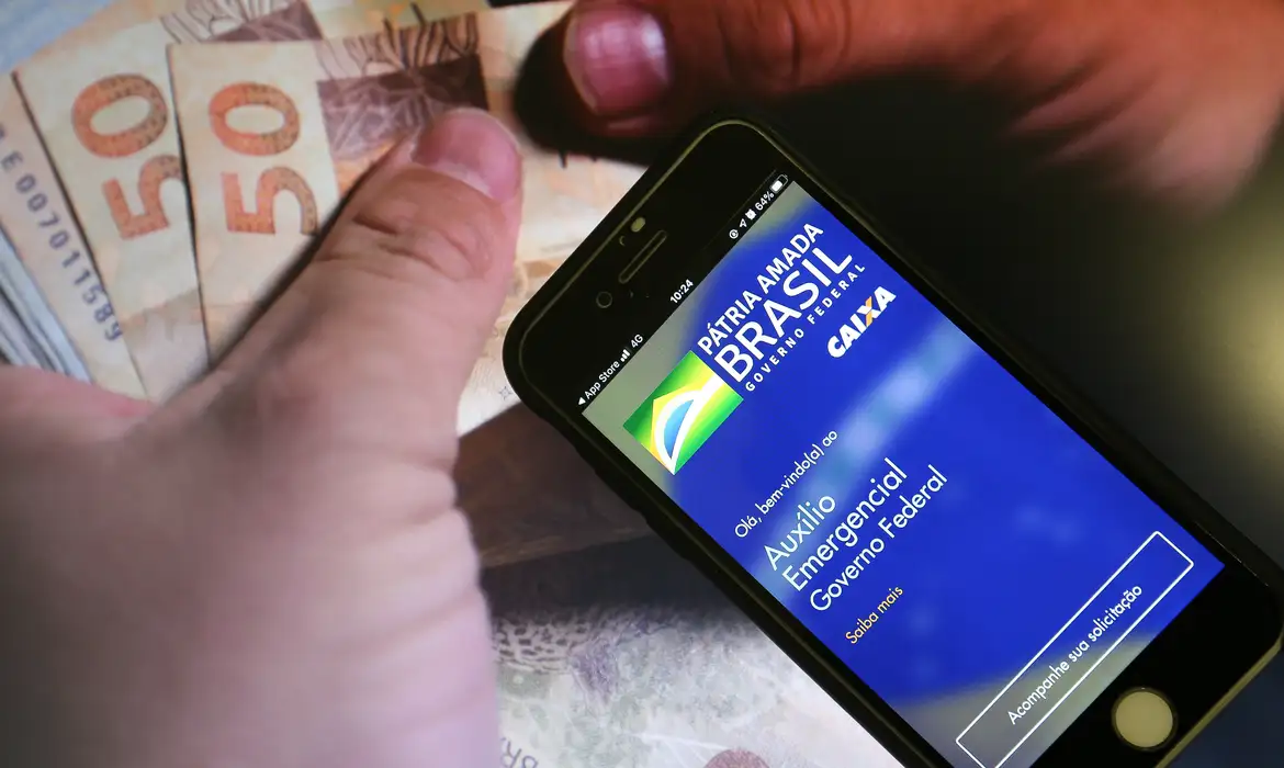 guia informativo auxilio emergencial governo Smartphone inclinado sobre mesa de escritório mostrando a interface do aplicativo oficial do Auxílio Emergencial em fundo azul vibrante.
